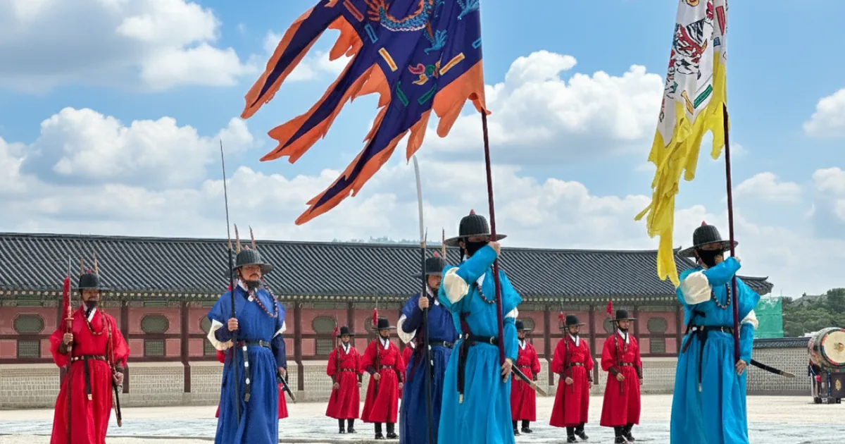 Gate guards in red and blue Joseon-era uniforms with tall yellow and blue banners during the changing ceremony at Gyeongbokgung