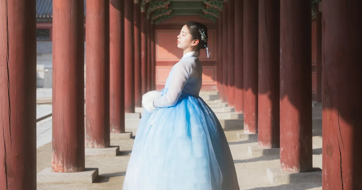A woman in a pale blue hanbok standing at the end of a long red-pillared palace corridor at Gyeongbokgung