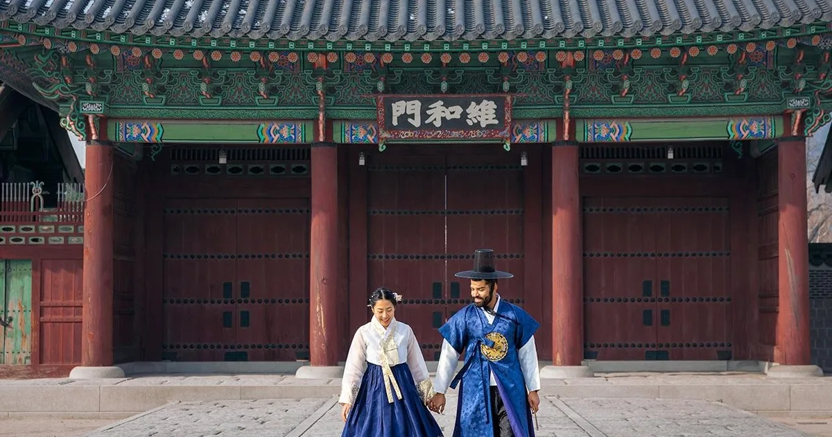 A couple in rented hanbok, she in a blue skirt and white jacket and he in a navy robe and black hat, walking out of a red palace gate at Gyeongbokgung